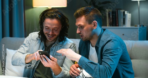 Handsome young Caucasian man showing something on smartphone to his best friend. Guys football fans putting bets at bookmakers on mobile phone in eveningt home on couch. photo