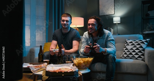 Two Caucasian friendly guys playing videogame with joystick while sitting in tension late in evening with snacks and beer in front of TV. Men having fun together with games. photo