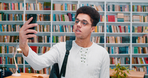 Handsome Latino young man in glasses standing in library and having videochat on smartphone. Male student talking via webcam on phone with books shelves on background.