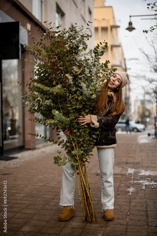 Girl in a dark jacket and jeans stands on a winter city street with a big green tree