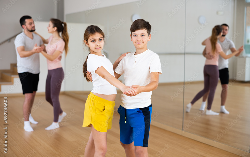 Pre-tee siblings practicing active dance in pair during family dance class, parents dancing on background