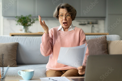 Senior woman sitting on couch at home shocked by bad unpleasant message or bank notice, feel frustrated by unexpected news in paper correspondence photo