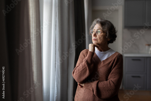 Retirement and depression. Upset thoughtful senior woman standing and looking at window at home, free space photo