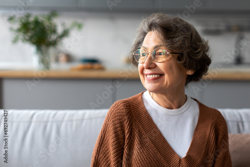Portrait of happy senior woman relaxing on comfortable couch at home in living room and looking away at free space, enjoying domestic pastime photo