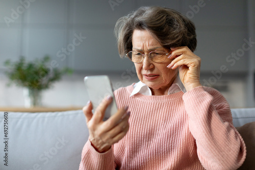 Confused senior woman in eyeglasses squinting eyes reading message on cellphone, having ophtalmic issue problems with vision sitting on couch