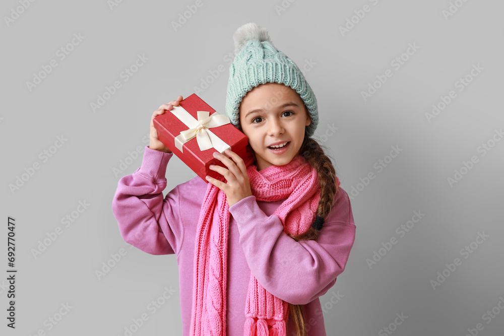 Cute little girl in warm hat and scarf with Christmas gift on grey background Cute little girl in warm hat and scarf with Christmas gift on grey background