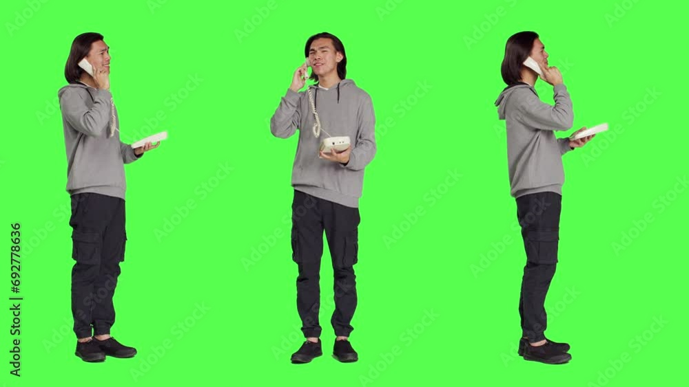 Asian guy talking on landline phone, answering call on telephone with cord while he stands against full body greenscreen. Young person using retro office phone for remote communication. Asian guy talking on landline phone, answering call on telephone with cord while he stands against full body greenscreen. Young person using retro office phone for remote communication.