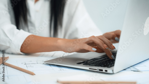 Close-up of young beautiful professional architect hands using laptop with blueprint and architectural document placed on table at modern office. Creative design concept. Focus on hand. Immaculate.