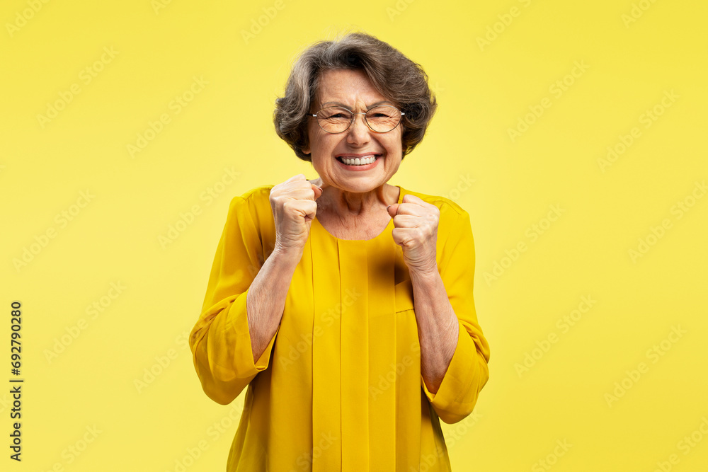 Funny emotional senior woman, happy modern grandmother holding hands celebration success isolated yellow background Funny emotional senior woman, happy modern grandmother holding hands celebration success isolated yellow background