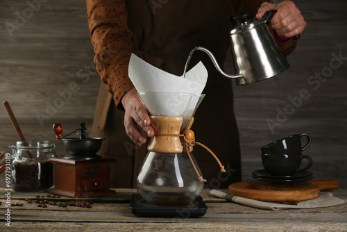 Woman pouring hot water into glass chemex coffeemaker with paper filter at wooden table, closeup photo