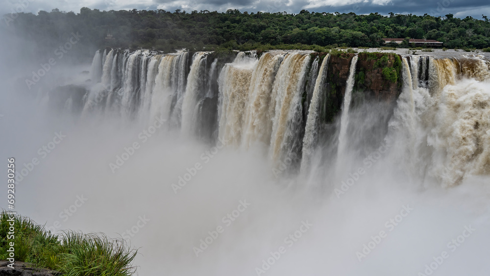 A tropical waterfall. Powerful foaming streams of water plunge into the abyss from the cliff. Splashes, thick fog in the air. Green grass in the foreground. Iguazu Falls. Argentina. A tropical waterfall. Powerful foaming streams of water plunge into the abyss from the cliff. Splashes, thick fog in the air. Green grass in the foreground. Iguazu Falls. Argentina.