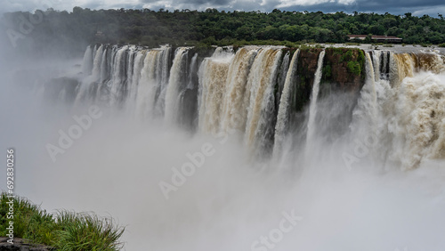 A tropical waterfall. Powerful foaming streams of water plunge into the abyss from the cliff. Splashes, thick fog in the air. Green grass in the foreground. Iguazu Falls. Argentina.