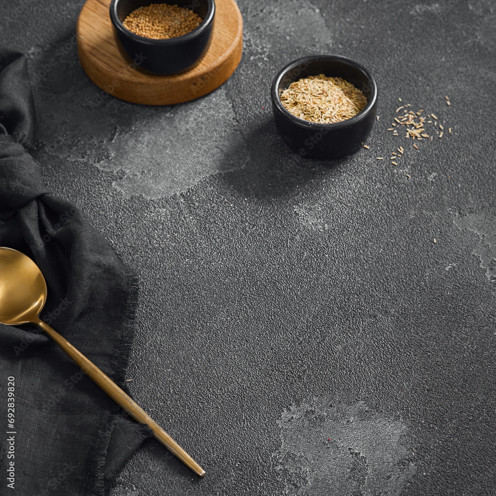 Stylish culinary setup with golden spoon and two bowls of spices on a textured grey background with a wooden base and black napkin Stylish culinary setup with golden spoon and two bowls of spices on a textured grey background with a wooden base and black napkin
