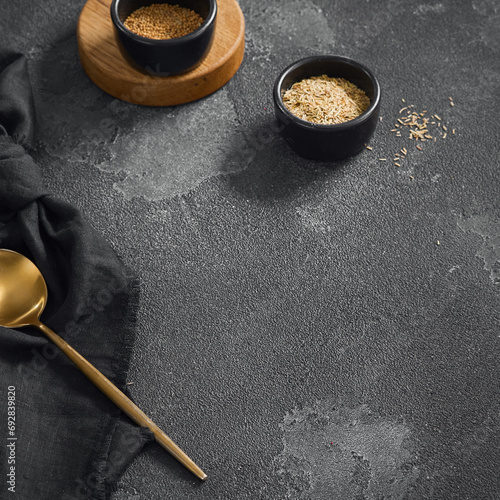 Stylish culinary setup with golden spoon and two bowls of spices on a textured grey background with a wooden base and black napkin