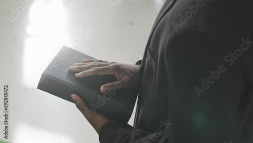 Cropped shot of unrecognizable Black male priest holding Bible while standing in sunlit church photo