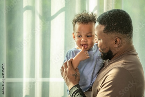 Happy african black parents dad father throw baby son on piggyback and neck riding teasing fun near window. Black baby son and daddy enjoy teasing kiss cheek and neck riding at window light photo