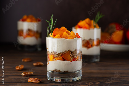 Vegan layered dessert in a glasses with persimmon, rosemary, pecan, whipped cream on a wooden table. Healthy food, sugar, gluten and lactose free.