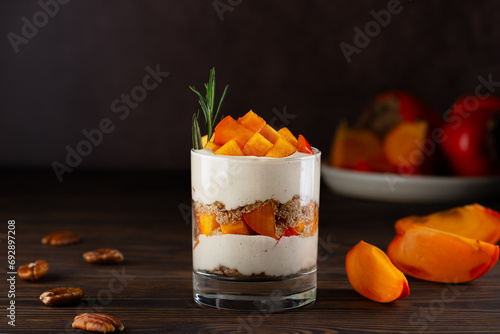 Vegan trifle in a glass with persimmon, rosemary, pecan, whipped cream and biscuit on a brown. Healthy food, sugar, gluten and lactose free.