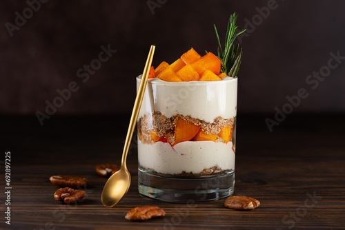 Vegan parfait in a glass with persimmon, rosemary, pecan, whipped cream and spoon on a brown. Healthy food, sugar, gluten and lactose free.