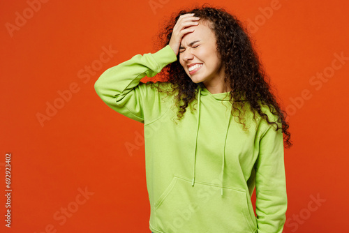 Young sad woman of African American ethnicity she wear green hoody casual clothes put hand on face facepalm epic fail mistaken omg gesture isolated on plain red orange background. Lifestyle concept.