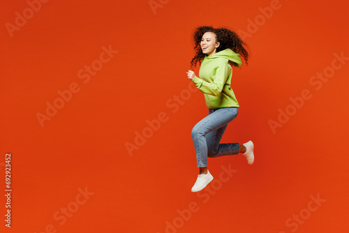 Full body side profile view young excited overjoyed woman of African American ethnicity wear green hoody casual clothes jump high run fast isolated on plain red orange background. Lifestyle concept.