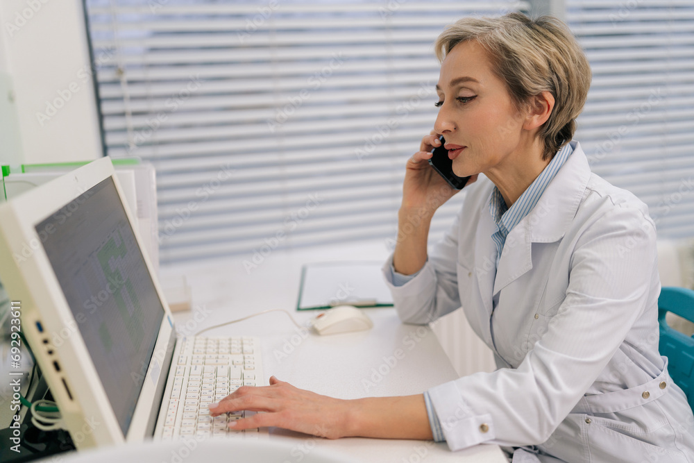 Side view of focused middle-aged woman general practitioner sit at desk in clinic office looking at laptop screen, discuss medical checkup results, talk to patient on cellphone work distantly. Side view of focused middle-aged woman general practitioner sit at desk in clinic office looking at laptop screen, discuss medical checkup results, talk to patient on cellphone work distantly.