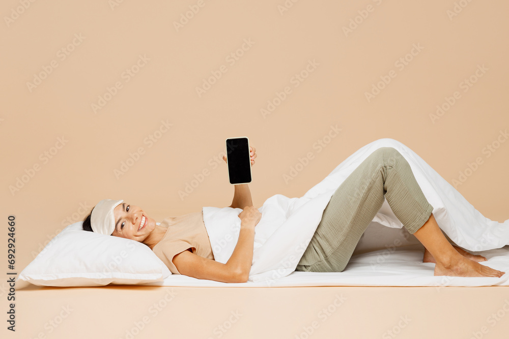Φωτογραφία Full body young Latin woman wears pyjamas jam sleep eye mask rest relax at home under duvet use blank screen mobile cell phone isolated on plain pastel beige background