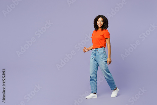 Full body side view smiling happy little kid teen girl of African American ethnicity she wear orange t-shirt walk go look camera isolated on plain pastel purple background Childhood lifestyle concept