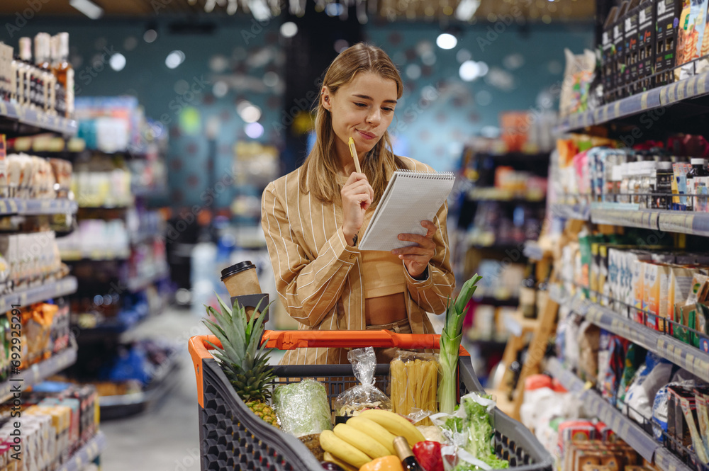 Young customer woman wears casual clothes write down in notebook list shopping at supermaket store grocery shop buy with trolley cart choose products in hypermarket Purchasing food gastronomy concept - obrazy, fototapety, plakaty 
