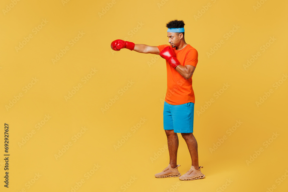 Full body young fitness trainer instructor sporty man sportsman wears orange t-shirt boxing red gloves punch spend time in home gym isolated on plain yellow background. Workout sport fit abs concept. Full body young fitness trainer instructor sporty man sportsman wears orange t-shirt boxing red gloves punch spend time in home gym isolated on plain yellow background. Workout sport fit abs concept.
