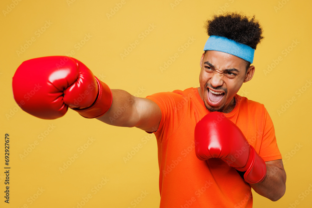 Young masculine fitness trainer instructor sporty man sportsman wear orange t-shirt boxing red gloves punch spend time in home gym isolated on plain yellow background. Workout sport fit abs concept. - obrazy, fototapety, plakaty 
