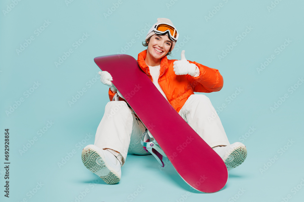 Φωτογραφία Full body young woman wear warm padded windbreaker jacket hat ski goggles mask sit with snowboard show thumb up travel rest spend weekend winter season in mountains isolated on plain blue background