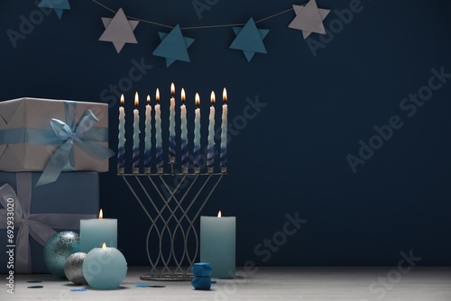 Hanukkah celebration. Menorah, burning candles, dreidels and gift boxes on white table against blue background, space for text photo