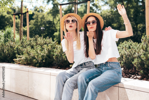 Two young beautiful smiling hipster female in trendy summer white t-shirt and jeans clothes. Carefree women posing in street. Positive models having fun. Cheerful and happy. In hat. Give air kiss