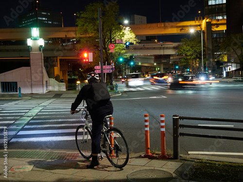 夜の東京の都心の街並みの交差点で信号待ちの一人の自転車を乗っている男性の後姿