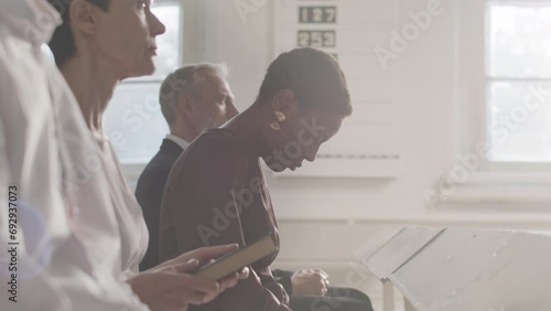 Lens flared waist up slowmo of Black woman and other diverse parishioners listening to pastor preaching sermon while sitting on bench together in church during religious service photo