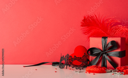 Romantic surprise awaits in discreet gift box adorned with sleek black bow. Explore pleasure with adult toys, furry handcuffs, feather tickler, and sprinkles. Side view photo, red background sets mood