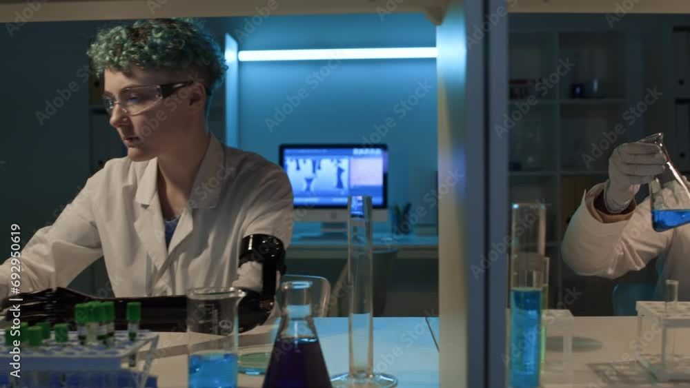 Medium shot of young female technician with prosthetic arm and mature Caucasian researcher sitting at desk in biochemistry lab, examining samples in test tubes, and typing on laptops Medium shot of young female technician with prosthetic arm and mature Caucasian researcher sitting at desk in biochemistry lab, examining samples in test tubes, and typing on laptops