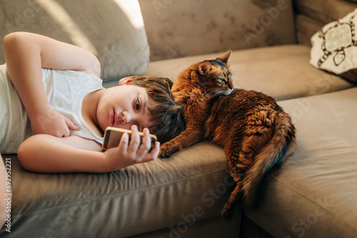Kid boy watching to his mobile phone lying on the couch sofa with his pet fluffy red Abyssinian cat together. Friendship with pets and support