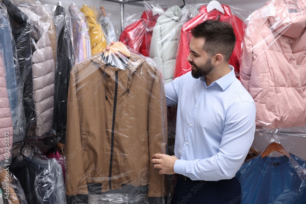 Dry-cleaning service. Happy worker holding hanger with jacket in plastic bag near other clothes indoors Dry-cleaning service. Happy worker holding hanger with jacket in plastic bag near other clothes indoors