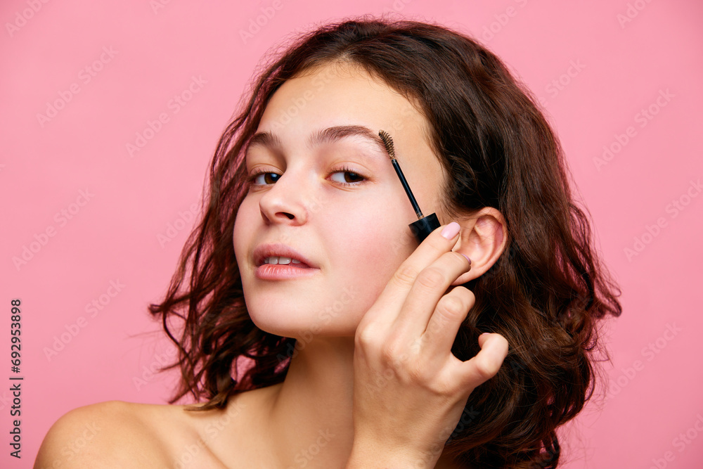 Pretty young brunette girl with short curly hair, doing makeup, applying eyebrows gel against pink background. Concept of natural female beauty, skin care, cosmetology and cosmetics Pretty young brunette girl with short curly hair, doing makeup, applying eyebrows gel against pink background. Concept of natural female beauty, skin care, cosmetology and cosmetics