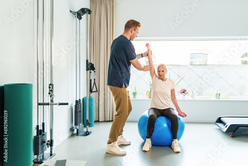 A focused senior woman sits on a blue fitness ball performing arm exercises with the guidance of her physiotherapist in a well-lit rehab gym.
