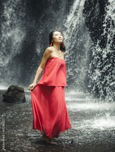 Asian woman posing near waterfall. Nature and environment. Water splashing. Travel lifestyle. Charming  woman wearing long red dress. Slim body. Copy space. Yeh Bulan waterfall, Bali