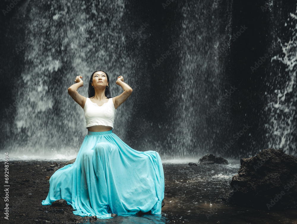 Beautiful Asian woman sitting near waterfall. Nature and environment. Water splashing. Travel lifestyle. Woman wearing long blue skirt. Slim body. Copy space. Yeh Bulan waterfall, Bali Beautiful Asian woman sitting near waterfall. Nature and environment. Water splashing. Travel lifestyle. Woman wearing long blue skirt. Slim body. Copy space. Yeh Bulan waterfall, Bali