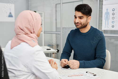Medium shot of smiling male Muslim patient listening to female doctor wearing headscarf during consultation in clinic photo