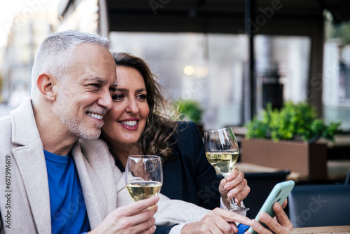 Happy senior couple sharing smartphone while drinking wine in city eating lunch at outdoors bar. Joyful mature man and woman using mobile phone together at a restaurant terrace.