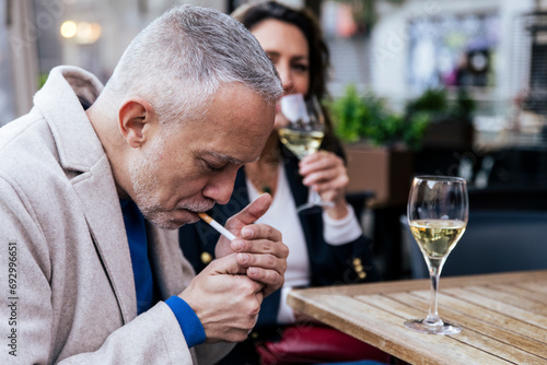 Senior man smoking a cigarette at a terrace while having a drink. Middle aged man smoking tobacco and drinking wine in a terrace with a woman. photo