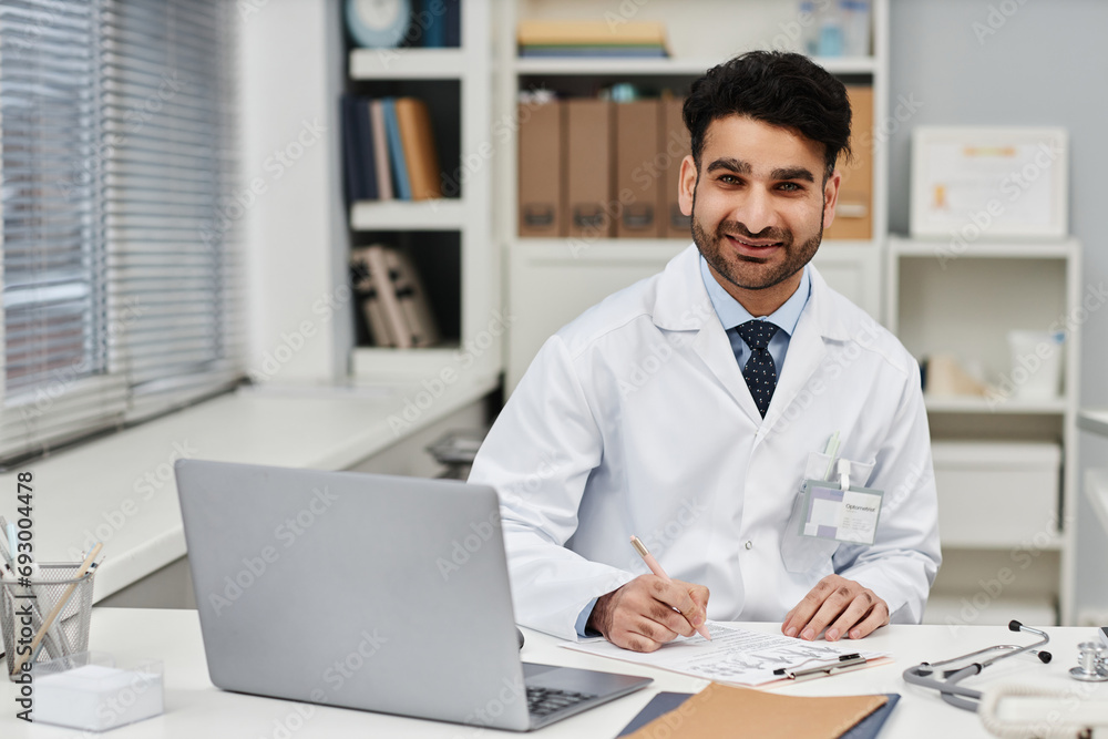 Fototapeta premium Portrait of cheerful Muslim man physician looking at camera while sitting at desk in clinic office