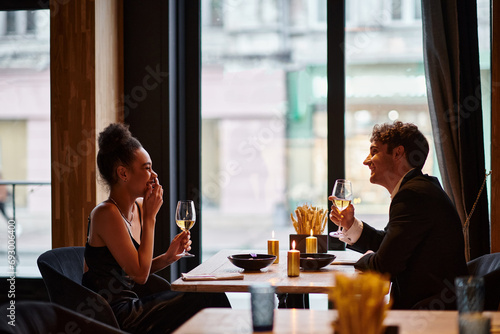 happy interracial couple in elegant attire holding glasses of wine during date in restaurant, laugh photo