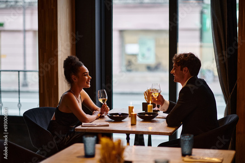 happy interracial couple in elegant attire holding glasses with wine during date in restaurant